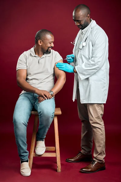 African american doctor giving covid vaccine to black man wearing surgical face mask. Full length healthy patient getting corona injection from physician against red studio background with copyspace.