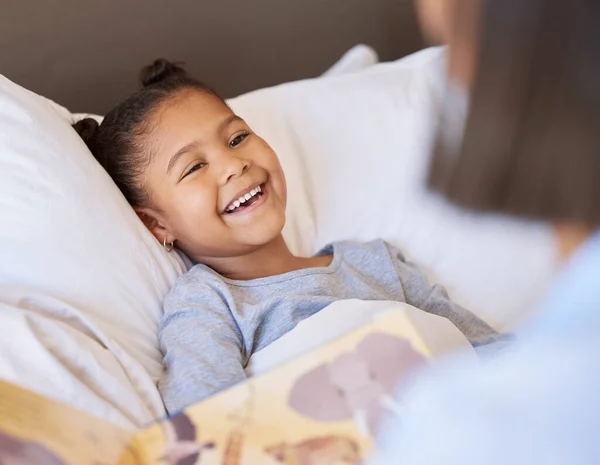 A cute little smiling mixed race girl laughing while lying in bed and enjoying story time. Young mother reading her adorable african American daughter a bedtime story while she relax in bed at home.
