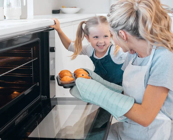 Happy mother and daughter removing muffins from the oven. Little girl looking at her parent holding a tray of muffins.Smiling caucasian girl making dessert muffins with her mother