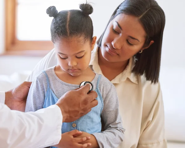 Sad little girl at doctors office. Sick girl sitting with mother while male paediatrician listen to chest heartbeat. Male doctor examining child with stethoscope. Mom holding kid during doctor visit.