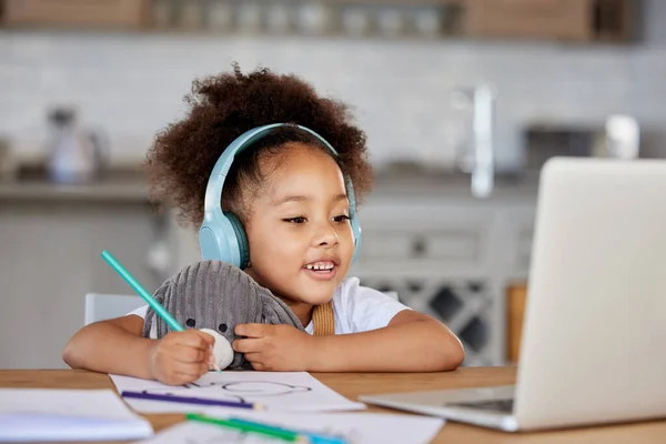 One mixed race preschool girl wearing headphones during video call with teacher on laptop for distance learning at home. Kid colouring with pencils during online virtual education class for homeschoo.