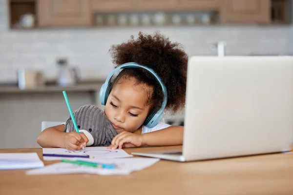 Adorable little girl wearing wireless headphone while learning online during video call with teacher. Young girl online with laptop while drawing and colouring with pencils during covid 19 pandemic.