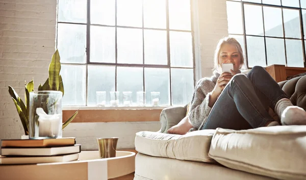 Living room, phone or relax and a woman in her home, sitting on the sofa with the sun shine through the window. Weekend, social media and communication with a young female relaxing in her house.