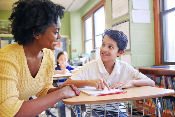 Learning, education and teacher help student with assignment in elementary school. Classroom support, scholarship and happy black woman or educator aid, explaining or helping male learner in class