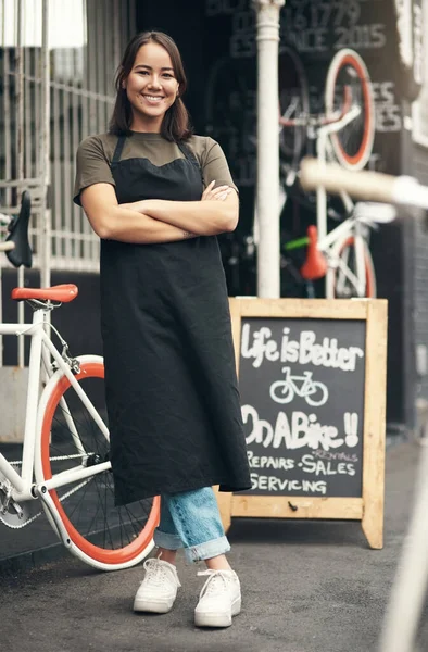 Lets get those training wheels off. Full length shot of an attractive young woman standing outside her bicycle shop with her arms folded