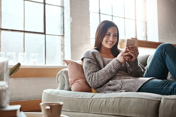 Phone, woman and relax on a sofa with app, social media and texting in her home, happy and smile. Smartphone, girl and chatting, online dating and internet, search and streaming in a living room.