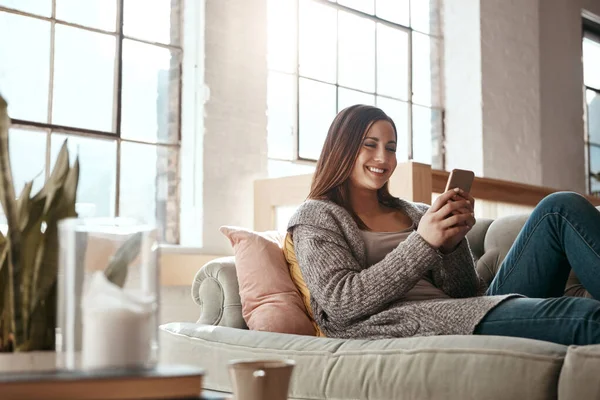 Happy, woman and phone for social media on a sofa, texting and chatting on dating app in her home. Girl, smartphone and text conversation in a living room, smile and relax while streaming and resting.