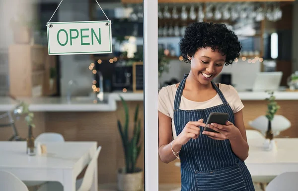 Digital tools support better business operations. a young woman using a cellphone while standing alongside an open sign in a cafe