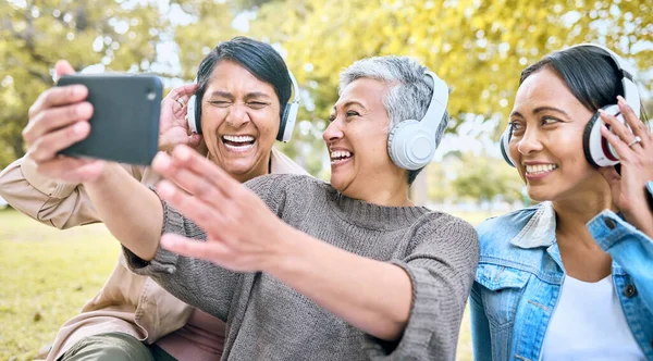 Senior woman, friends and phone laughing with headphones listening to music or watching comedy movie in the park. Group of happy elderly women enjoying funny series or 5G connection on smartphone.