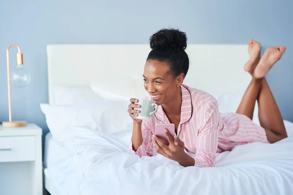 That sounds like a pretty good way to spend my day. an attractive young woman drinking coffee while using her cellphone in bed at home