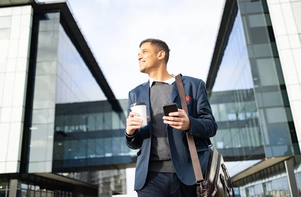 Businessman outside building with phone, coffee and bag, happy waiting for online taxi service after work. Office, business and success, man with smile holding 5g smartphone standing on city street