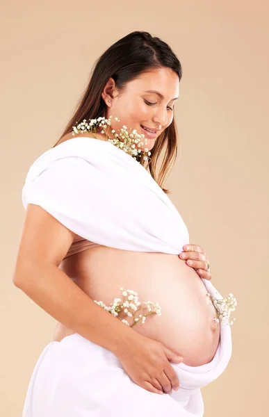 Pregnant, mother and flowers on stomach, healthcare and excited for birth, smile and girl on brown studio background. Pregnancy, woman and mama with tummy, plants and nurture for wellness and growth.