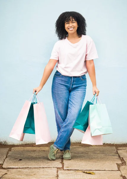 Shopping, retail and portrait of a woman with bags from a sale on a blue city wall in Greece. Fashion, product and girl shopper carrying content from a shop, boutique or store downtown with a smile.