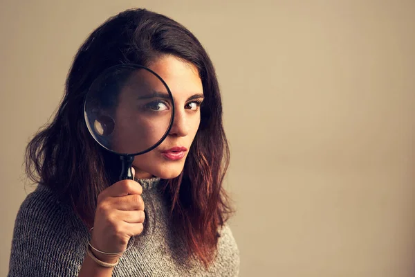 Yes, I see you there. Studio portrait of a young woman looking through a magnifying glass against a brown background