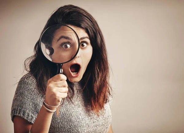 When you finally find what youve been stalking for. Studio portrait of a young woman looking shocked while using a magnifying glass against a brown background