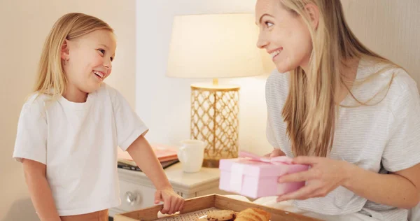 Breakfast, child and mama with a gift on mothers day in the morning to celebrate her mommy at home. Smile, Love and happy girl giving parent a present box and a croissant with cookies in a bedroom.