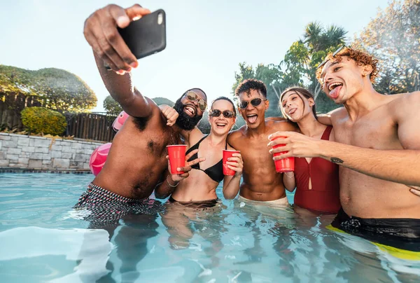Selfie, beer and friends at pool party having fun on new years. Swimming celebration, water event and group of happy people with tongue out, peace sign and taking pictures for social media in summer