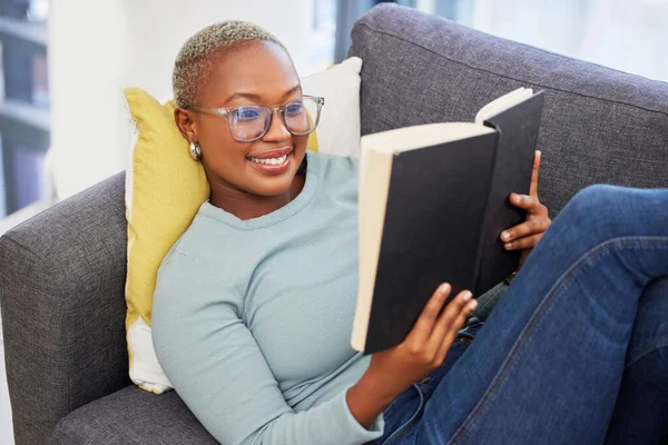 Black woman, relax smile and reading book on sofa in living room for calm peace, happiness and studying in home. Young african girl, happy student and relaxing with notebook or university textbook.