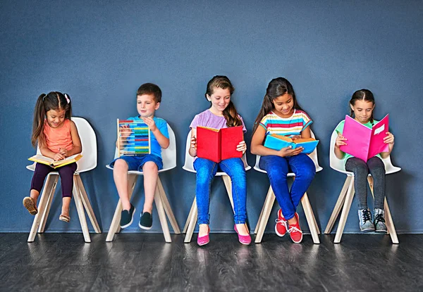 Learn something new everyday. Studio shot of a group of kids sitting on chairs and reading books against a blue background