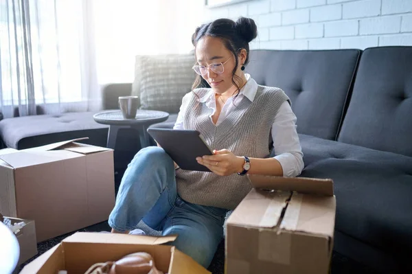 Real estate, tablet and Asian woman moving into new home while planning or calculating mortgage online. Relocation boxes, technology and female property owner or remote worker in living room of house.