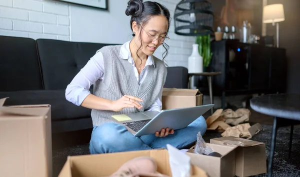 Real estate, laptop and Asian woman moving into new home while planning or calculating mortgage online. Relocation boxes, computer and female property owner or remote worker in living room of house