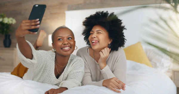 Happy, friends and selfie with phone in bed with a smile, relax and funny during a luxury vacation in a hotel room. African american women having fun with social media post on a 5g mobile smartphone.