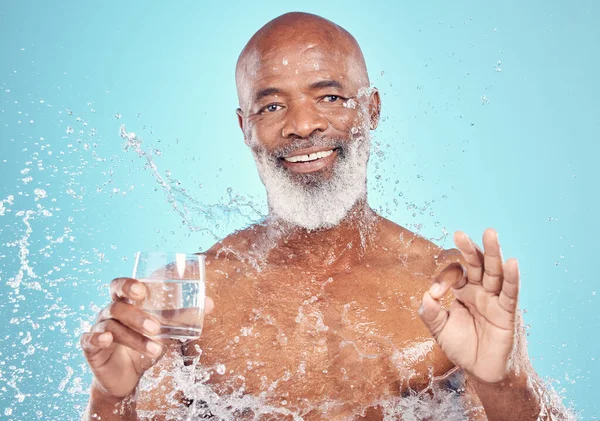 Splash, portrait and elderly man in a studio with a glass of water and a ok hand gesture. Health, wellness and happy senior African male with sign language by a blue background with mockup space.