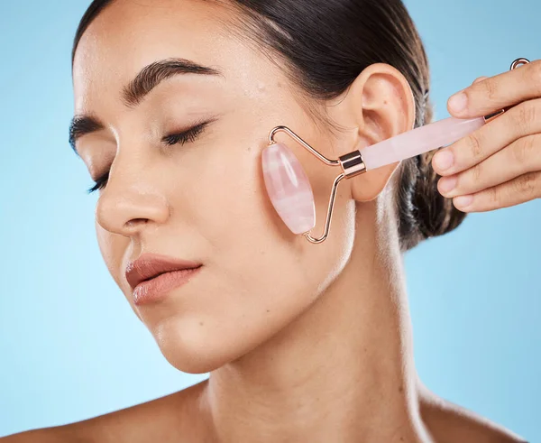 Skin care, face roller and beauty woman with facial massage for dermatology, cosmetic and wellness. Young aesthetic model person for natural rose quartz spa product to relax on blue background.