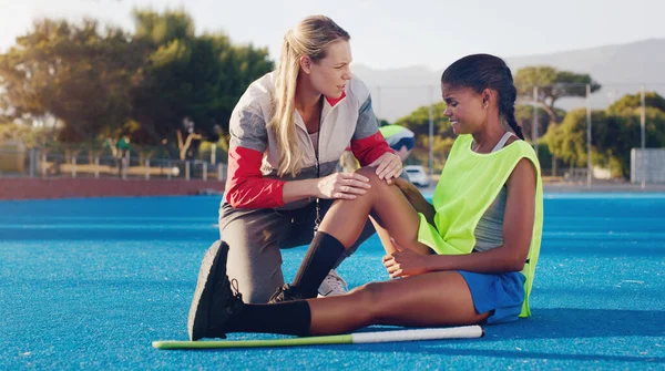 Women, hockey coach and knee injury girl on field together with support, solidarity or care at sport training. Sports woman, coaching or help at workout for joint pain at outdoor exercise with mentor.