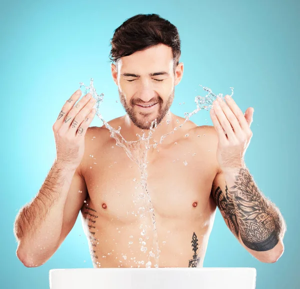 Water splash, sink and skincare of a man cleaning face for wellness, self care and dermatology. Liquid, model ad blue background in a studio with a young man with tattoo doing treatment for beauty.