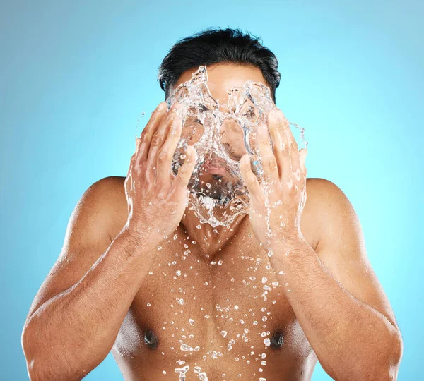 Man, face and water splash in studio with skincare, wellness and grooming on blue background. Cleaning, beauty and moisture by Mexican model relax with luxury, routine and facial while isolated.