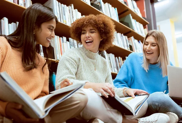 Friends, students in library with books for education, university group of women, funny and studying together. Laughter, diversity and young, research for paper or exam with motivation and reading.
