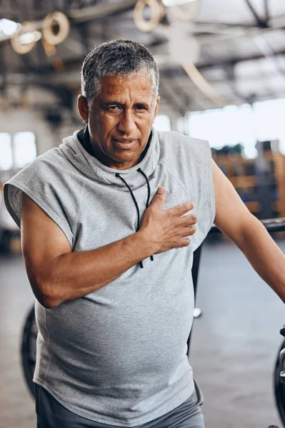 Fitness, gym and old man with pain in chest, medical emergency during workout at sports studio. Exercise, health and wellness, stress and tired senior person with hand on heart muscle while training