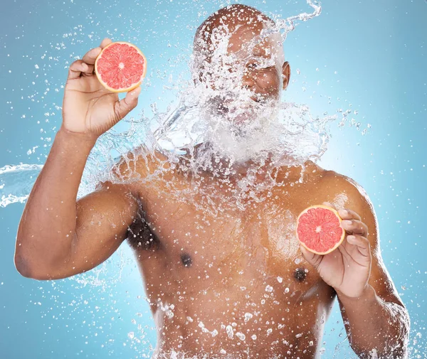 Black man, water splash and fruit for skincare, grapefruit or vitamin C and hydration against a blue studio background. Happy African American male smiling and holding food for health and wellness.