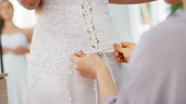 Friends helping woman with wedding dress knot in a room or boutique for marriage ready, fitting or fashion with corset lace detail and hands. Bride with mirror reflection getting ready for love event.