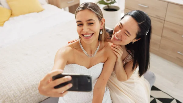 Bride, bridesmaid and selfie with phone, wedding and ring with a smile to post on social media. Happy woman and asian friend excited for marriage celebration with fashion white dress in a bedroom.