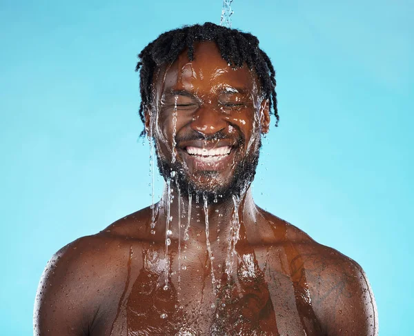 Water drops, skincare and black man isolated on blue background for face cleaning, body shower and smile. Strong, beauty model or person, facial glow in studio headshot washing or dermatology hygiene.