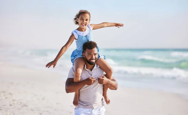 Happy father carrying his daughter on his shoulders while walking along the beach. Adorable little girl stretching out her arms and pretending to fly while on holiday and enjoying family time with da.