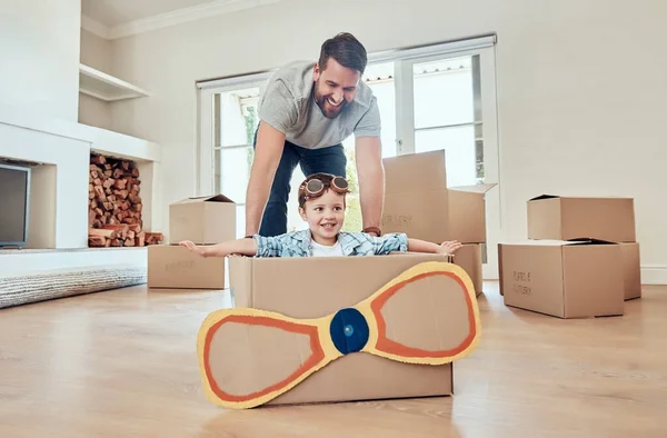 Happy single father pushing his little kid in homemade aeroplane cardboard box at home. Adorable little boy sitting in makeshift plane and playing with single parent in a living room in their new hom.