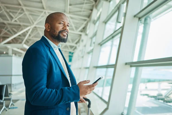 Black man, phone and thinking at airport window for business travel, trip or communication waiting for flight. African American male with smile contemplating schedule or plain times on smartphone.