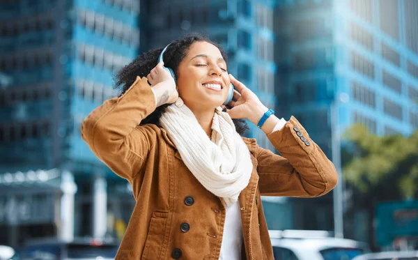 Black woman, headphones and listening to music in city for travel, motivation and happy mindset. Young person on an urban street with buildings background while streaming podcast or audio outdoor.