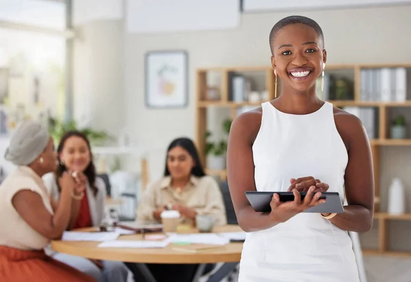 Smiling african american business woman using a digital tablet while colleagues sit behind her in office. Ambitious and happy black professional standing and holding technology while browsing schedul.