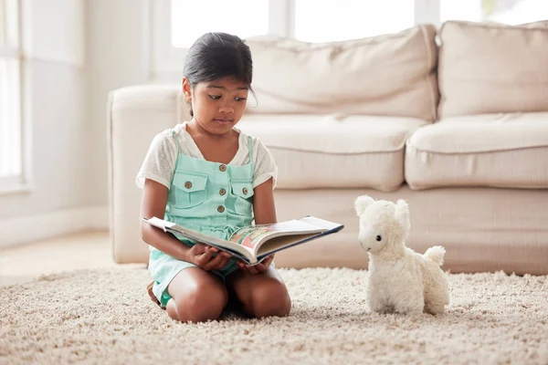 Girl playing pretend and reading a book to her toy while sitting on the floor at home and learning.