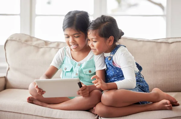 Two little girls playing online with a digital tablet, looking happy and content on the sofa at home.