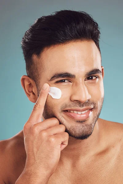 Portrait of one smiling young indian man applying moisturiser lotion to his face while grooming against a blue studio background. Handsome guy using sunscreen with spf for uv protection. Rubbing faci.