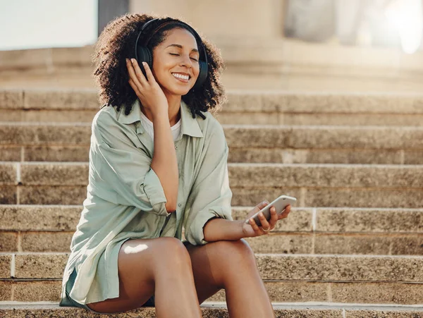 Black woman outdoor, mobile phone and headphone for music, travel and 5g network for audio streaming in city. Happy in Miami with peace, listen to podcast and relax on steps with internet connection.