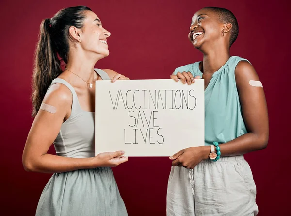 Covid vaccinated African american and mixed race women showing and holding poster. Two people isolated on red studio background with copyspace. Showing plaster on arm and promoting corona vaccine.