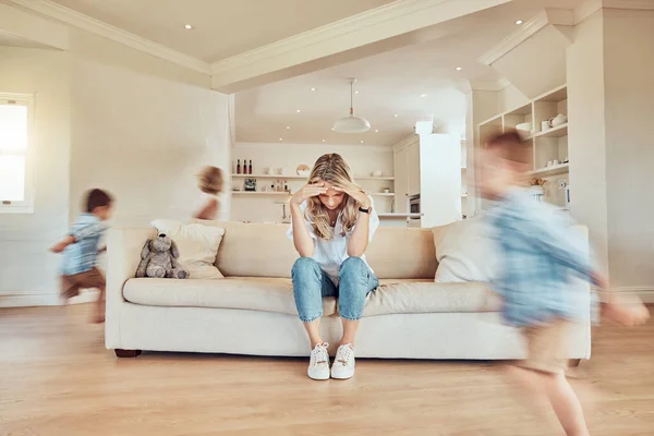 Stressed mother sitting on the sofa while her adhd children run around the living room. Hyperactive son and daughter giving single parent a headache. Active siblings chasing each other in the house.