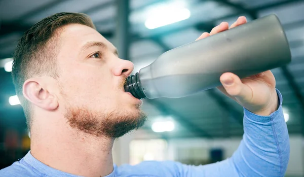 Gym, fitness and man drinking water during training, exercise and intense cardio, serious and thirsty on blurred background. Hydration, athletic and male with bottle during workout at sports center.