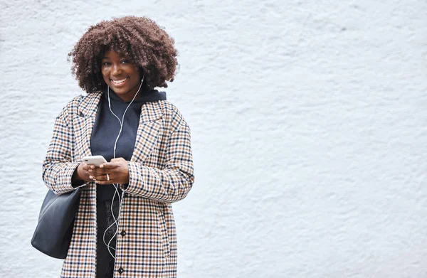 Black woman, phone and portrait smile listening to music with earphones in fashion and afro hairstyle on mockup. Happy African American female holding smartphone for audio track on a wall background.